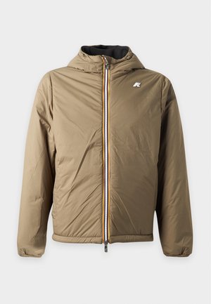 Jeune homme portant un sweat à capuche bleu marine zippé sur un polo noir, un jean large bleu clair et des baskets blanches avec des détails noirs, debout devant un fond uni.