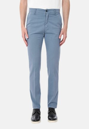 Uomo che indossa pantaloni slim fit azzurri chiari, camicia bianca e scarpe nere con suole bianche, in piedi davanti a uno sfondo neutro.