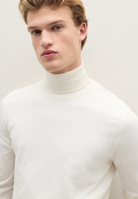 Junger Mann mit hellem Haar, der einen weißen Rollkragenpullover trägt und vor einem schlichten, hellen Hintergrund nach vorne schaut.