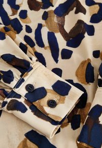 Tissu beige avec des taches abstraites irrégulières bleu marine et marron, montrant un gros plan d'un poignet de manche longue avec deux boutons noirs.