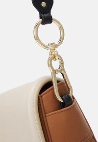 Primo piano di una borsa in pelle beige e marrone con una tracolla nera attaccata da un anello e un gancio in metallo dorato.