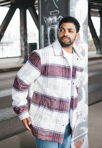 Bărbat îmbrăcat într-o jachetă albă și maroniu în carouri și blugi stând sprijinit de un pilon acoperit cu graffiti sub un pod.