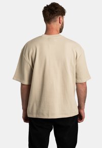 Beige žebrované tričko s kulatým výstřihem, krátkým rukávem a volným střihem, zobrazené z zadní strany na bílém pozadí.