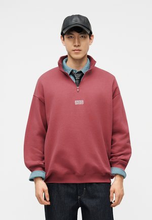 Ung mand iført en sort læderbaseballkasket, bordeaux kvart-zip sweatshirt over en denimskjorte og sort slips, stående mod en hvid baggrund.