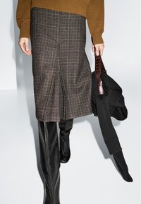 Osoba ubrana w brązowy sweter, spódnicę w kratę oraz czarne, skórzane kozaki do kolan, trzymająca złożony czarny dzianinowy sweter z brązowym skórzanym paskiem.