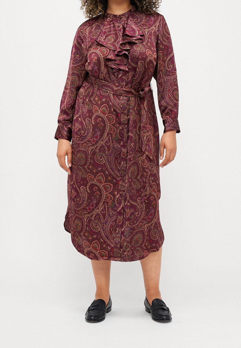 Lungo abito paisley bordeaux con colletto a balze a strati, maniche lunghe, vita con cintura e texture liscia. Indossato con mocassini neri.