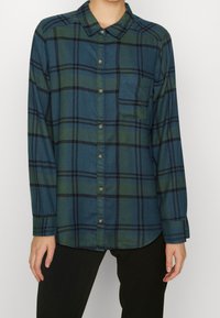 Koszula w kratę zapinana na guziki w ciemnozielonym i niebieskim kolorze z kieszonką na piersi, z długimi rękawami i kołnierzykiem w kształcie punktu. Miękka faktura materiału.