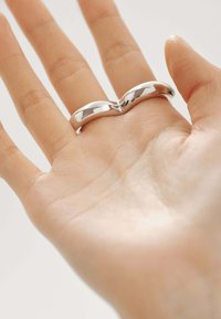 Anillo plateado elegante con un diseño en forma de ola, que presenta un acabado liso y pulido, reposando sobre una palma con textura de piel visible.