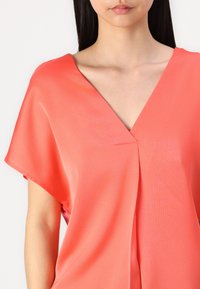 Femme portant un chemisier corail à col en V avec manches courtes, montrant le cou et le haut du torse sur un fond blanc uni.