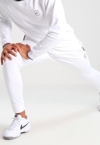 White athletic tracksuit with a long-sleeve top and tapered pants, featuring a small logo on the chest, paired with white Nike shoes.