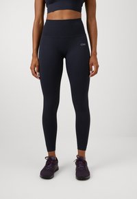 Granatowe legginsy z prążkowanej tkaniny z wysokim stanem, z logo na biodrze, w połączeniu z ciemnofioletowymi butami sportowymi. Gładki materiał i dopasowany krój.