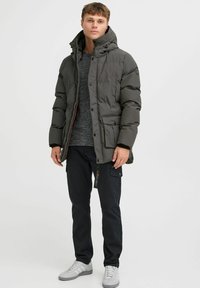 Chaqueta acolchada de color verde oscuro con capucha, que presenta un cierre de cremallera y botones, un ajuste oversized, y múltiples bolsillos frontales. Suéter gris texturizado debajo, pantalones negros y zapatillas blancas.