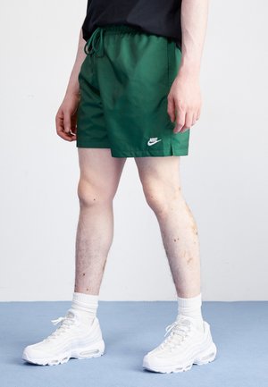 Grüne Nike Badehosen mit elastischem Bund und Kordelzug, die ein weißes Logo am linken Bein tragen. Getragen mit weißen Sneakers und Socken.