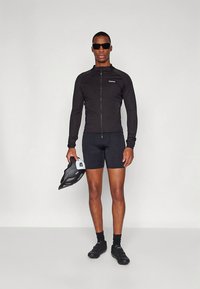 Črna kolesarska jakna z zadrgo, črne kratke hlače in črni kolesarski čevlji. Model drži črno čelado in nosi temna sončna očala.