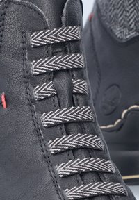 Bottine en cuir noir avec des lacets en chevron gris, finition texturée et couture d'accent rouge près de la zone des oeillets.