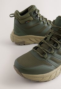 Žalios spalvos žygio batai su aukštu kulkšnies daliu, tekstūruotu tinkleliu ir guminiu viršumi, storu smėlio spalvos padu bei išryškintomis raištelėmis tvirtesniam prisitaikymui.