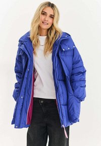 Modrá prošívaná bunda s lesklým povrchem, vysokým límcem, velkými kapsami a cvoky, nošená přes bílou košili.