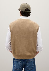 Maglione beige a maniche scoperte lavorato a maglia con una texture a coste, indossato sopra una camicia bianca, abbinato a jeans in denim blu e un cappello texturizzato.