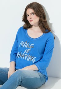 Nuori nainen, jolla on laineikkaat ruskeat hiukset, ja hänellä on korvassa renkaat, päällä sininen paita, jossa lukee "Make it Amazing", istuu valkoista seinää vasten.