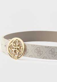 Vaalean beige kuvioitu kangasvyö, jossa on pyöreä kultasävyinen logovirike. Remmissä on hienovarainen logokuviointi ja sileä sisäpinta.