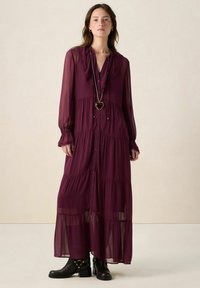 Robe longue bordeaux avec un col à volants, un devant boutonné et une jupe à plusieurs niveaux. Fabriquée en tissu léger, associée à des bottines noires.