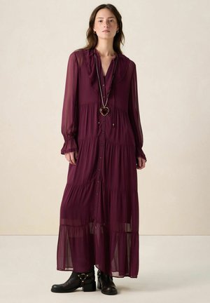 Langes burgunderrotes Kleid mit einem Rüschensaum am Ausschnitt, Knopfleiste vorne und mehrstufigem Rock. Hergestellt aus leichtem Stoff, kombiniert mit schwarzen Stiefeletten.