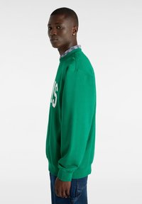 Sudadera verde con cuello redondo, con letras blancas. Hecha de una tela suave, con puños de canalé y un ajuste relajado. Llevada sobre una camisa de cuadros.