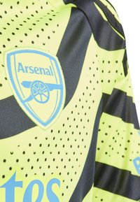 Camiseta de fútbol amarilla y negra con el logo de Arsenal, textura punteada, diseño angular y mangas cortas con rayas horizontales.