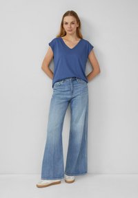 Eine Frau trägt ein blaues ärmelloses Oberteil mit V-Ausschnitt, weit geschnittene hellblaue Jeans und weiße Sneaker. Sie steht vor einer schlichten grauen Wand.