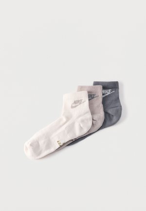 Trois chaussettes de cheville Nike pliées en blanc, beige et gris, empilées côte à côte sur un fond uni.