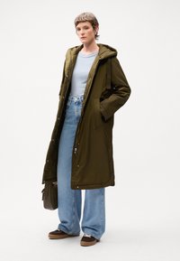 Olijfgroene lange parka met een capuchon, rits en drukknoopsluiting, zijzakken, gecombineerd met een lichtblauw shirt en wijde denim jeans.