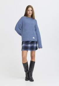 Maglione in pile blu con vestibilità oversize, abbinato a una minigonna scozzese blu e nera, completato da stivali neri alti fino al ginocchio.