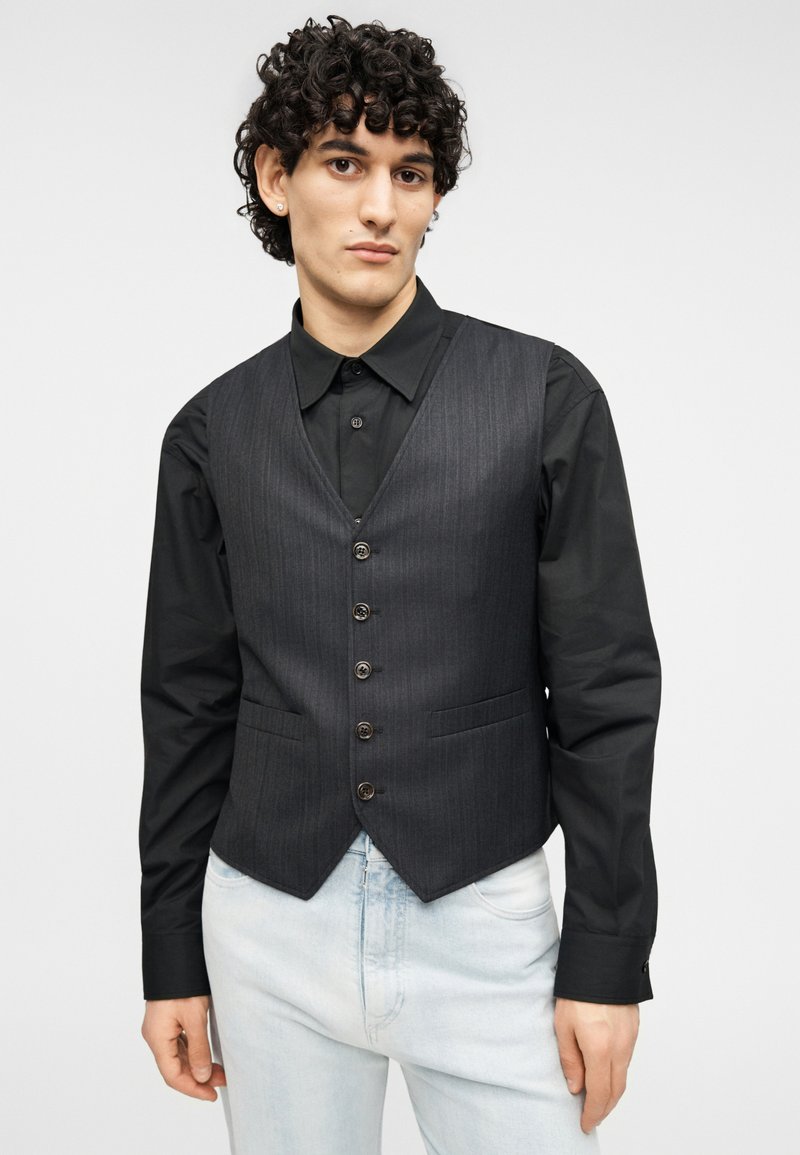 Gilet gris à fines rayures avec un col en V, comportant six boutons et deux poches avant, porté sur une chemise noire à manches longues et un jean denim clair.