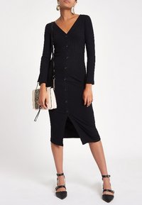 Robe noire côtelée avec un col en V, manches longues, fermeture boutonnée à l'avant et une fente mi-longue. Associée à un petit sac  à main beige et des talons noirs.