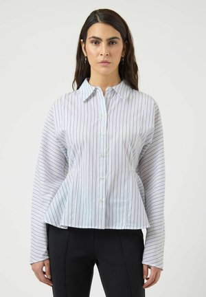 Femme aux cheveux foncés portant une chemise blanche à boutons avec de fines rayures verticales noires et un pantalon noir, debout devant un fond uni.