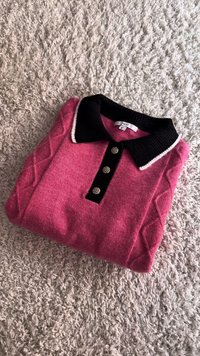 Maglione polo lavorato a maglia rosa con colletto a coste nero, tre bottoni decorativi e motivi a rombo sulle maniche. Piegato ordinatamente.