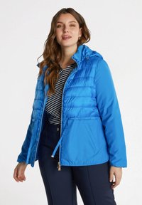 Chaqueta acolchada azul con capucha, que presenta costuras horizontales, cierre de cremallera en la parte delantera y cordón ajustable en la cintura. Llevada sobre una camiseta a rayas.