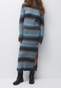 Robe tricotée à manches longues avec des rayures horizontales en gris et bleu, dotée d'une fente sur le côté et de poignets côtelés. Portée avec des bottines noires.