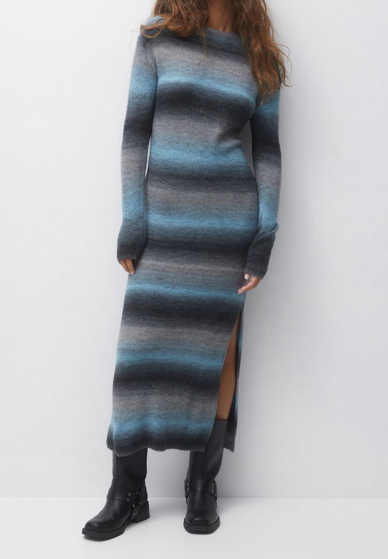 Robe tricotée à manches longues avec des rayures horizontales en gris et bleu, dotée d'une fente sur le côté et de poignets côtelés. Portée avec des bottines noires.