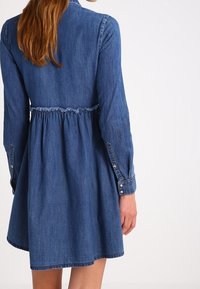 Denimmekko, jossa on kapea vyötärö, pitkät hihat, kaulus ja nappikoristeet. Ominaisuuksiin kuuluu liesattu hame ja vaaleansininen väri, jossa on teksturoitu kangas.