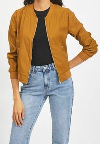 Hořčicová bavlněná bomber bunda se zipem na přední straně, žebrovanými manžetami a lemem. V kombinaci s džínami světle modré barvy s vysokým pasem. Hladký textura.