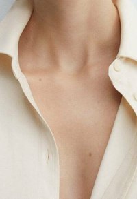 Camisa de color crema con cuello que presenta una botonadura y tela suave. Primer plano del escote muestra una textura lisa y un ligero brillo.