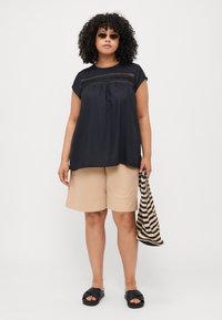 Top corto negro con detalles de encaje, combinado con pantalones cortos beige. La modelo sostiene un bolso tejido a rayas y lleva sandalias negras de tiras.