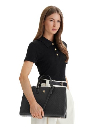 Žena v černém polo tričku s krátkým rukávem a bílých kalhotách drží černou texturovanou kabelku se zlatým logem a dvěma držadly.