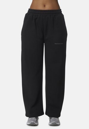 Persona che indossa pantaloni della tuta neri larghi con vita elastica e scarpe da ginnastica grigie, visibile dalla vita ai piedi su uno sfondo uniforme.