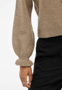 Manche de pull en tricot beige avec poignet élastique côtelé sur un pantalon noir, main avec des bagues en argent reposant à côté de la cuisse.