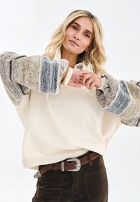 Kremowa, oversize'owa bluza z fakturowanymi, wzorzystymi rękawami w odcieniach szarości i brązu, zestawiona z brązowym paskiem z ozdobną srebrną klamrą.