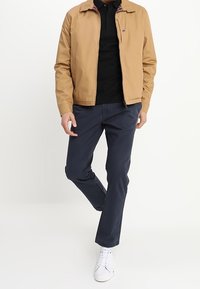 Mies, joka on pukeutunut vaaleanruskeaan takkiin mustan polo-paidan, tummansinisten housujen ja valkoisten lenkkarien kanssa, seisoo yhdellä jalalla taivutettuna.