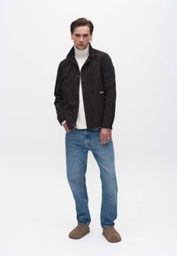 Schwarze Button-up-Jacke mit Kragen, getragen über einem weißen Rollkragenpullover, kombiniert mit hellblauen Jeans und braunen Slippern.