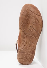 Hnědá kožená podrážka sandálu s texturovaným povrchem, zdobená rytinami a logem. Mírná křivka; minimální design vzorku.